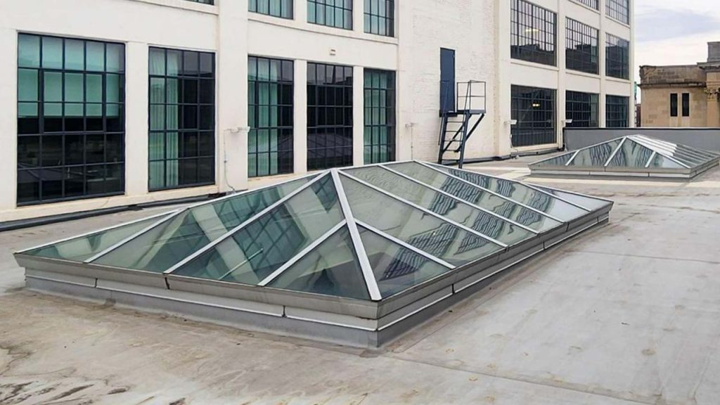Extended pyramid-shaped glass skylights on a flat industrial rooftop, framed by metal, with large windows and a metal staircase in the background, creating a modern feel.