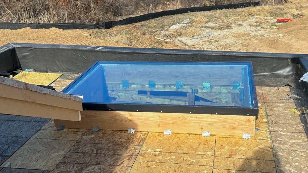 A transparent rectangular glass skylight set in a wooden frame on a construction site roof. Surrounding area shows dry grass and bare soil, suggesting a work in progress.