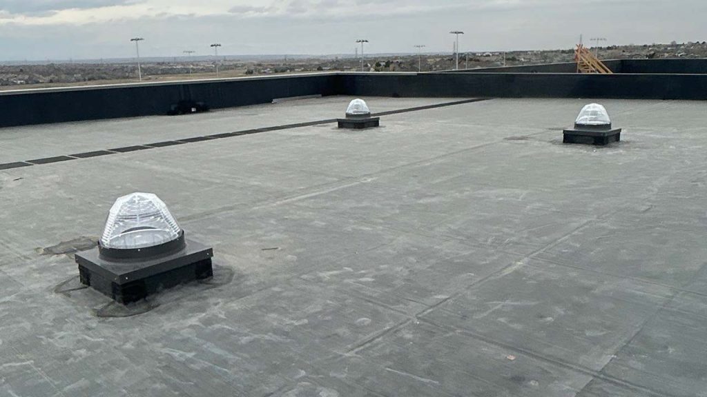 Flat rooftop with three evenly spaced VELUX TCC 022 Sun Tunnel skylights on a cloudy day. The surface is gray, with cityscape visible in the distant background.