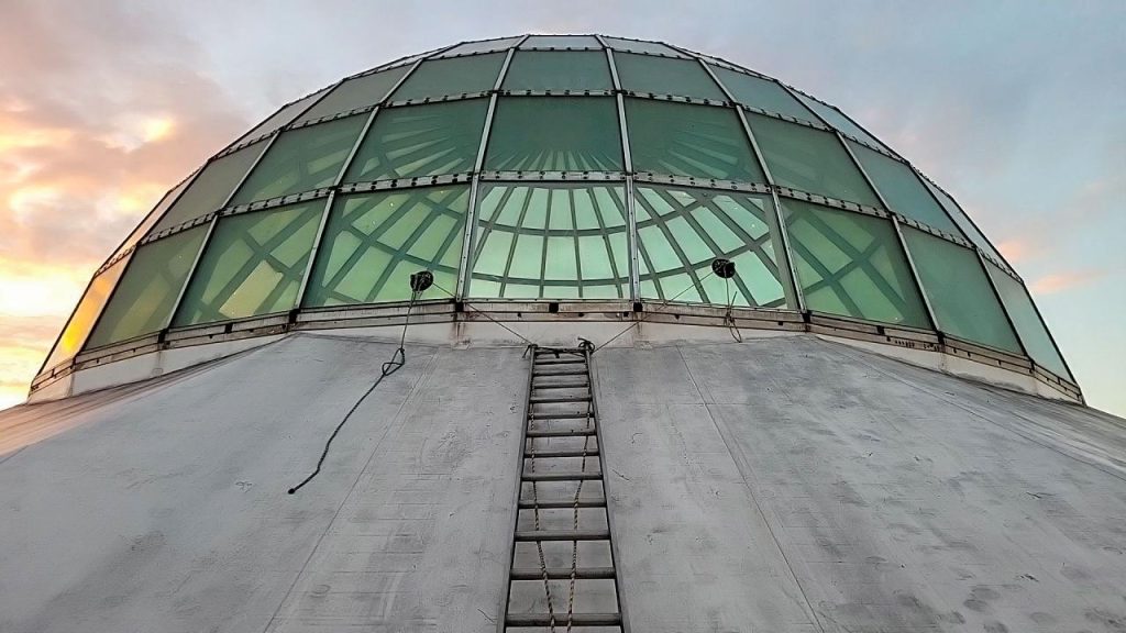 A green glass-domed skylight against a pastel sunset sky. A ladder leads up the silver dome, creating a symmetry that feels industrial and futuristic.