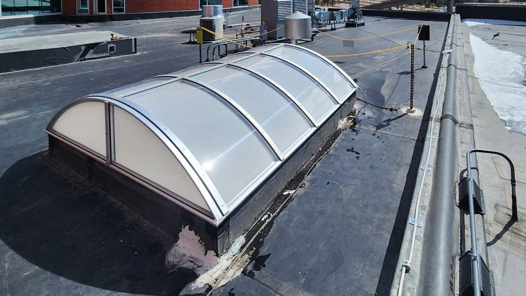 A curved, translucent barrel vault skylight on a flat rooftop is surrounded by industrial equipment and a safety rail. The scene is well-lit, indicating a clear day.