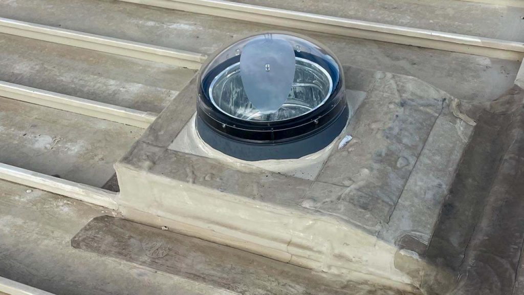 A rooftop skylight tube with a clear, dome-shaped cover set amidst a flat, weathered surface. The structure allows natural light into the building.