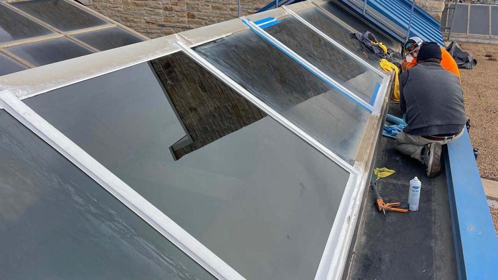 Worker in safety gear repairs a rooftop skylight on a modern building. Tools and spray can nearby, with clear skies reflected in the glass panels.