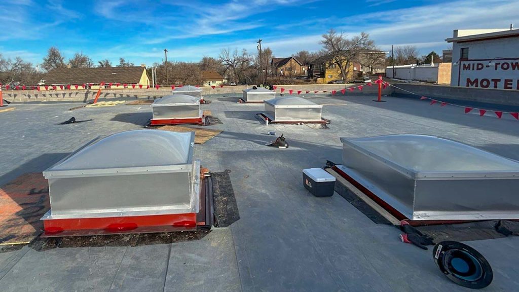 A flat rooftop with multiple rectangular skylights and red safety flags. The sky is clear and blue, with buildings and trees in the background.