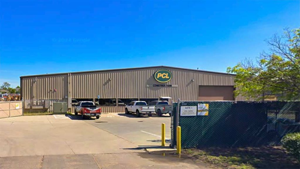 Large industrial warehouse with a "PCL Construction" sign. Three parked trucks in front, blue sky above, and a chain-link fence with a gate in the foreground.