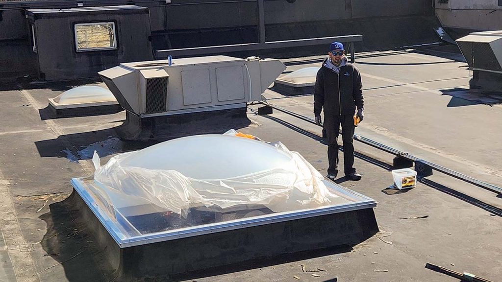 A worker in safety gear stands on a flat rooftop beside a newly installed skylight, partially covered with plastic. The scene conveys a sense of industrial work and modern construction.