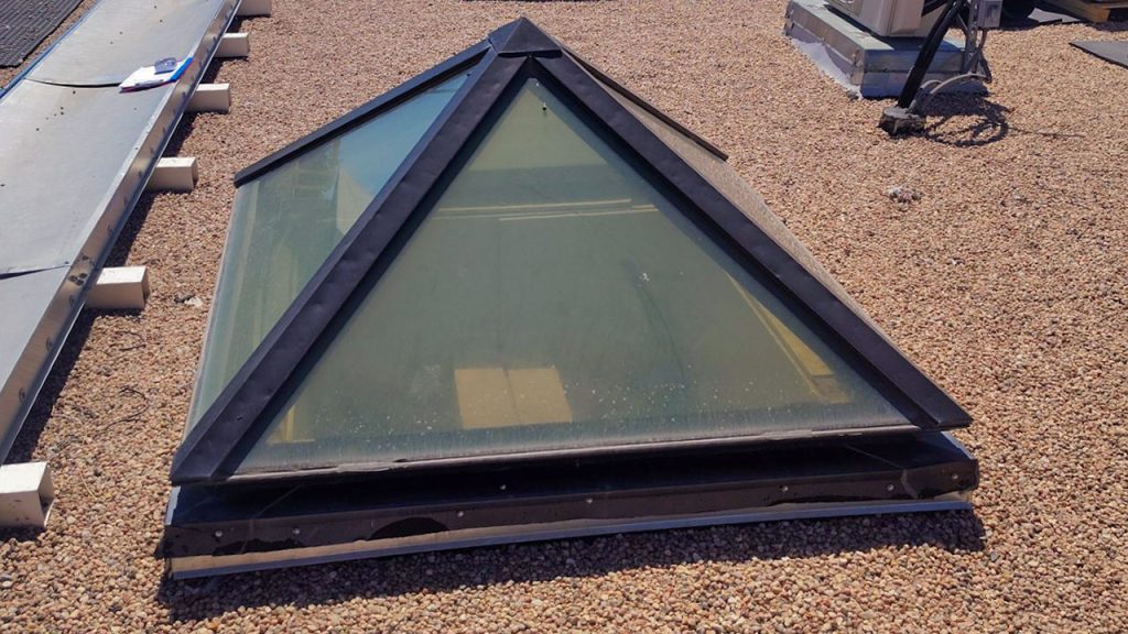 Pyramid-shaped skylight on a gravel-covered rooftop, surrounded by utility structures, creating a stark, industrial atmosphere under clear daylight.