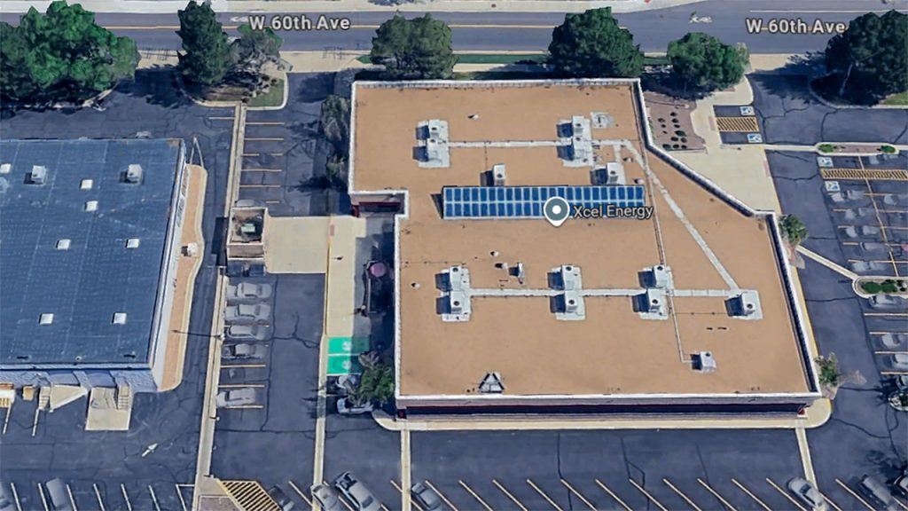 Aerial view of a flat-roofed building labeled "Xcel Energy," featuring glass ridge skylight in the center, adjacent parking lots, and surrounding greenery.