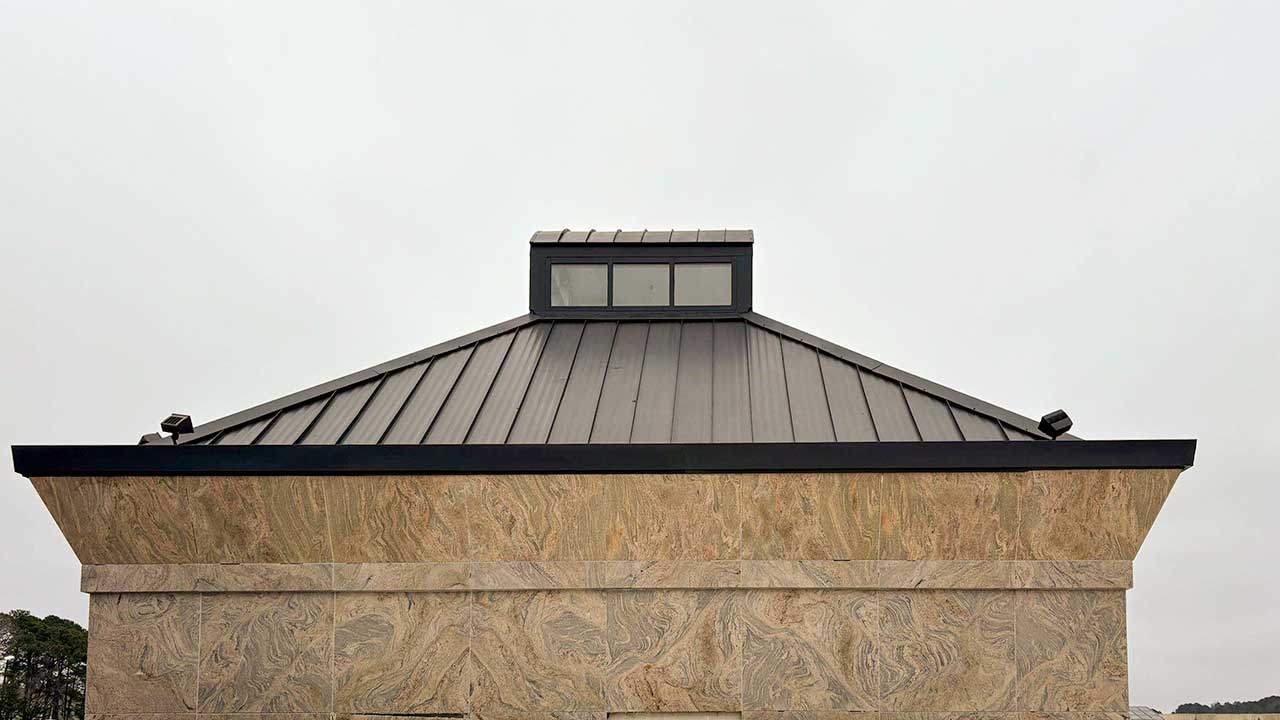 A modern building with a textured stone wall and a sloped metal roof topped by rectangular windows in cupola. The overcast sky creates a neutral, calm tone.