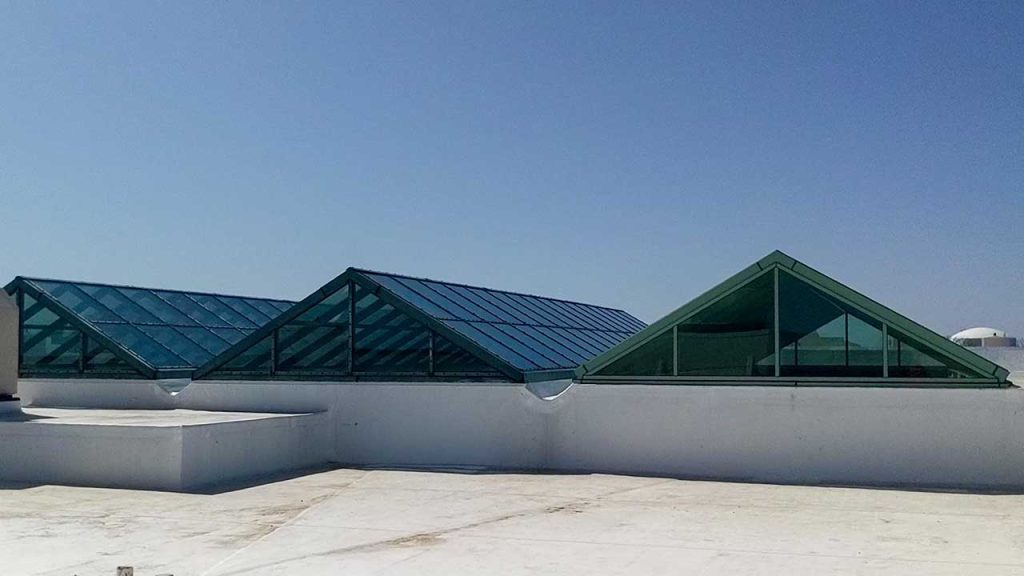 Triangular glass skylights sit atop a white rooftop under a clear blue sky, allowing natural light to enter the building and creating a modern, bright ambiance.
