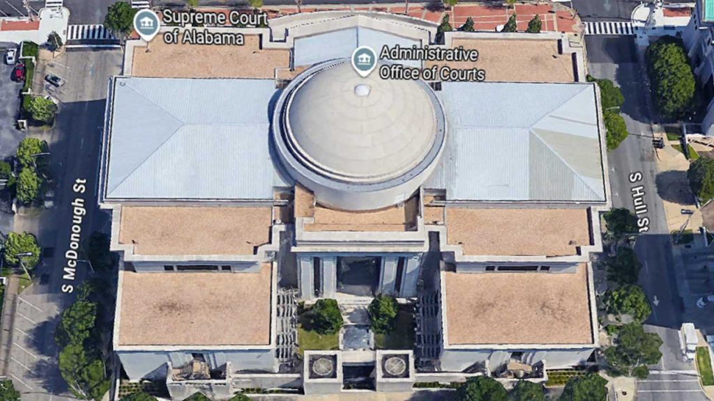 Aerial view of a large, neoclassical building with a prominent dome at the center, housing the Supreme Court of Alabama and Administrative Office. Streets lined with trees surround the building.