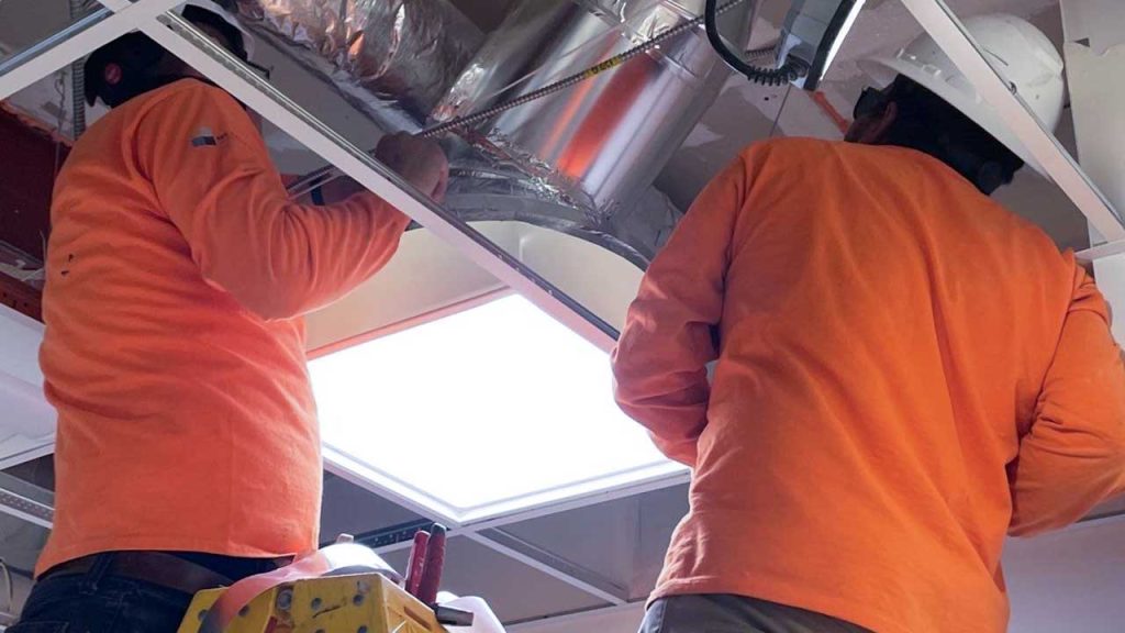 Two workers in orange shirts and hard hats work on installing a Velux Sub Tunnel in an industrial setting. The scene conveys focus and teamwork.