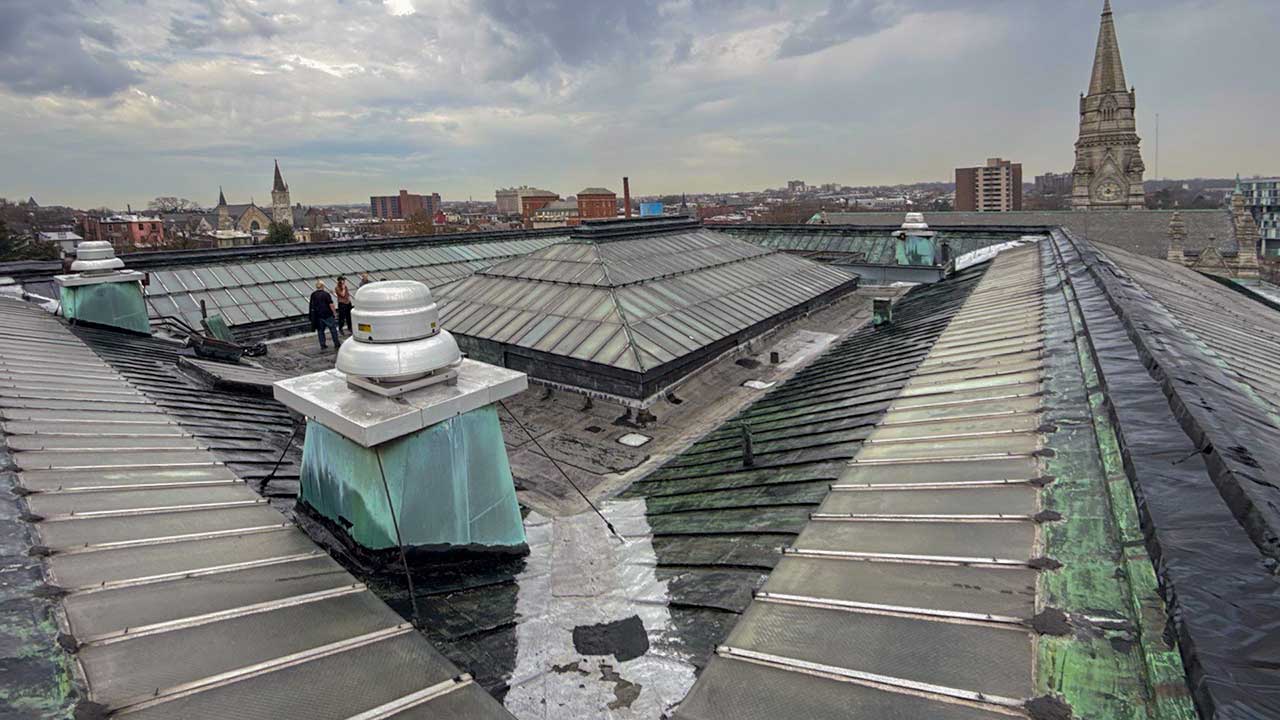 Rooftop view featuring geometric glass skylights and weathered green copper. Two people stand on the roof, with a cityscape and church spire in the background. Cloudy sky adds a moody tone.