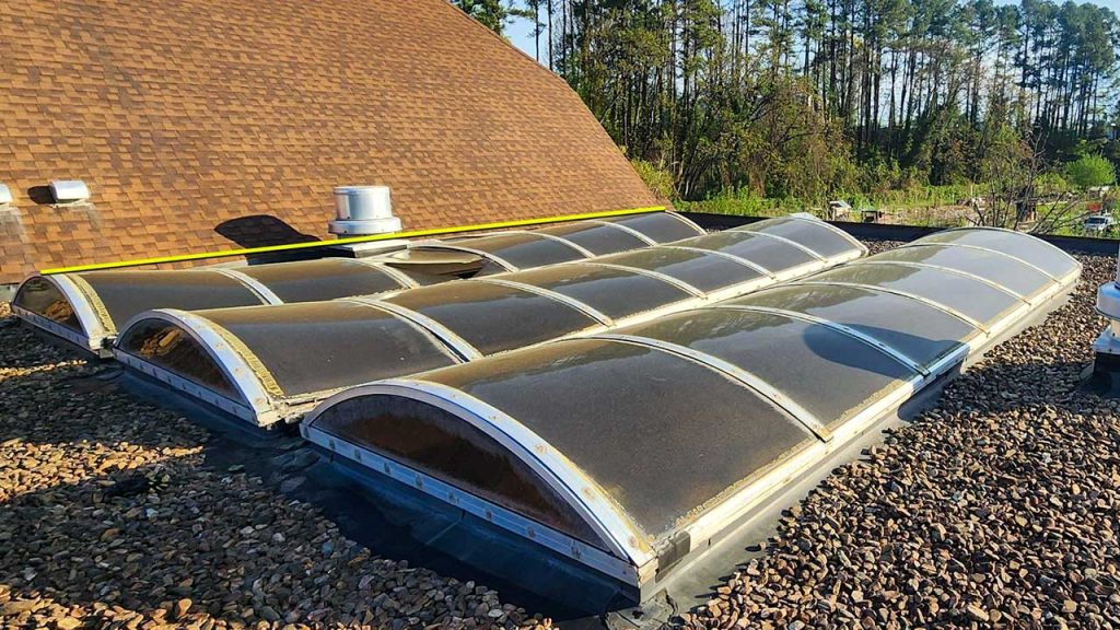 Three barrel vault skylights on a rooftop are surrounded by small rocks. Sunlight casts shadows, and trees are visible in the background.