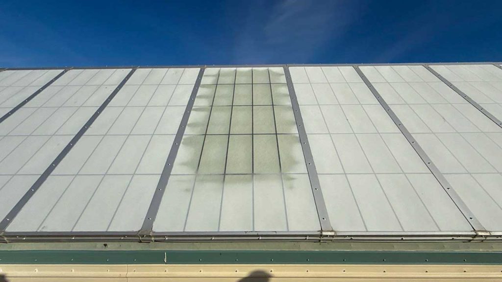 A building roof with a translucent, fiberglass skylight with a failed section that is discolored with green residue, under a clear blue sky conveying a serene tone.
