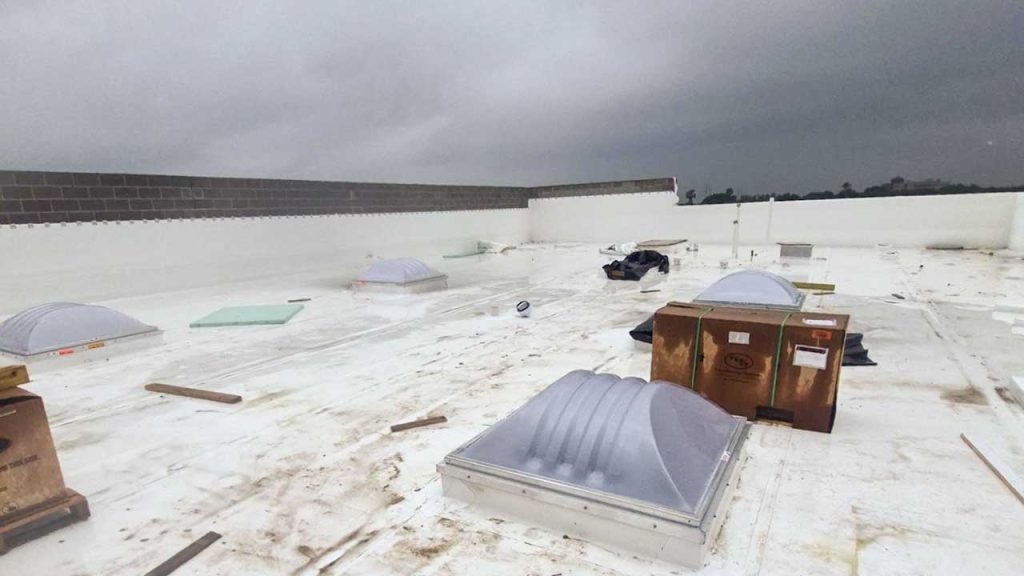 Flat rooftop with multiple white dome skylights is pictured under overcast skies. Debris is scattered, suggesting construction or maintenance work.