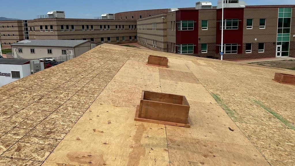 Flat wooden roof under construction with rectangular openings, set against a modern brick building. The sky is clear, creating a calm atmosphere.