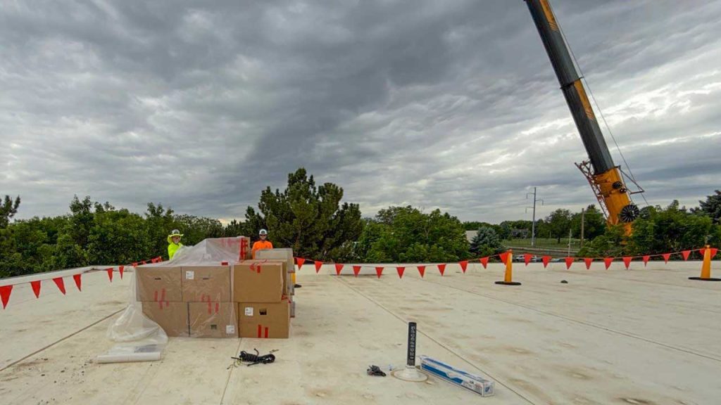 Two workers in safety gear stand on a roof near large boxes, surrounded by red safety flags. A crane is visible against a cloudy sky.