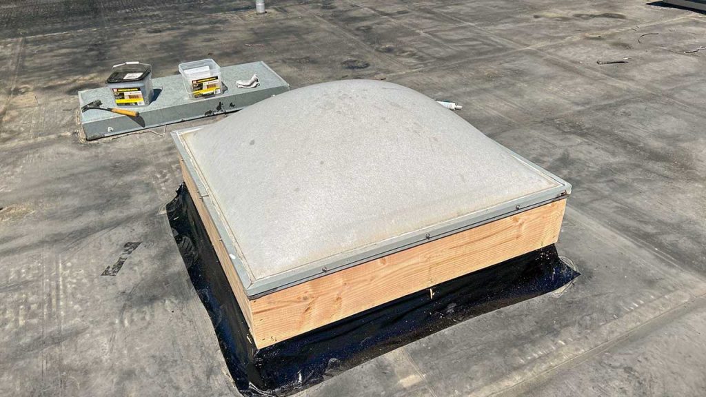 A flat rooftop with a dome-shaped skylight surrounded by a wooden frame. Nearby are construction tools and materials, conveying ongoing maintenance.