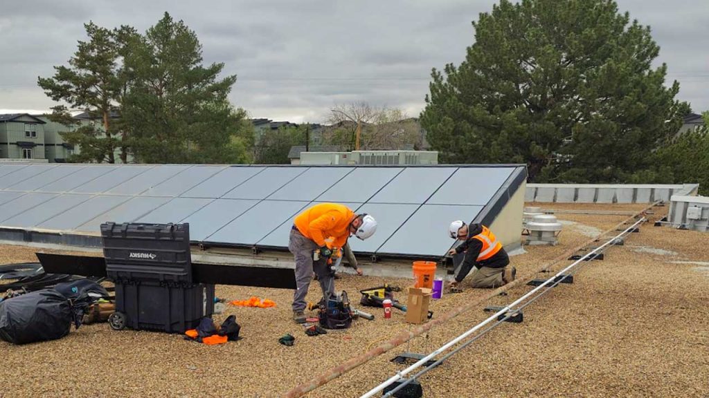 Two workers in orange safety vests and repairing a skylight on a gravel rooftop, surrounded by tools and equipment. Cloudy sky, trees nearby.