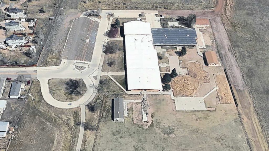 Aerial view of a large, L-shaped Arp Elementary School complex with solar panels on the roof. Surrounded by parking, small buildings, and open green fields.