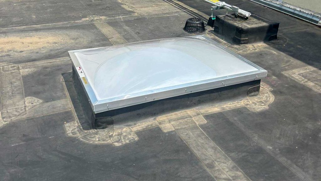 Rounded rooftop skylight with a clear dome set on a flat, dark roof. Nearby, a small black vent and maintenance equipment are visible. Bright, sunny atmosphere.