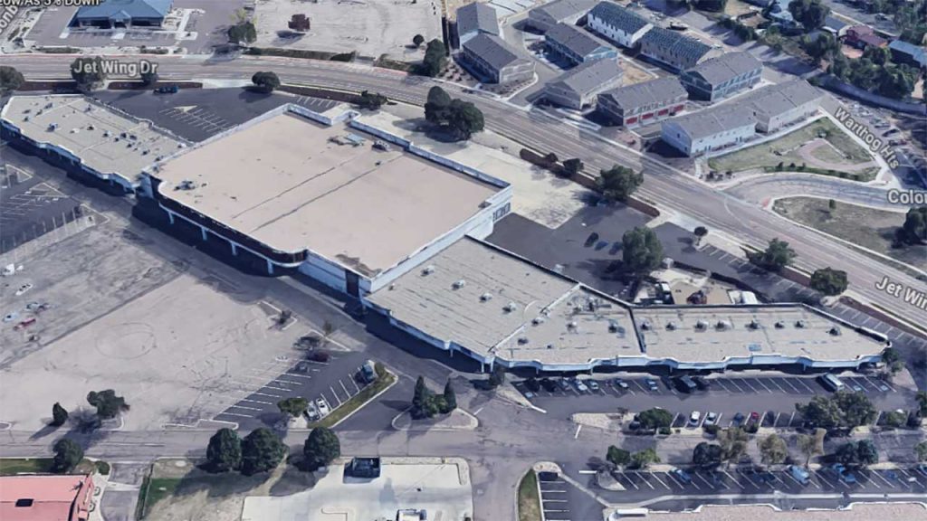Aerial view of a large, L-shaped shopping center with a flat, tan roof, surrounded by parking lots and trees. Nearby streets include Jet Wing Drive.