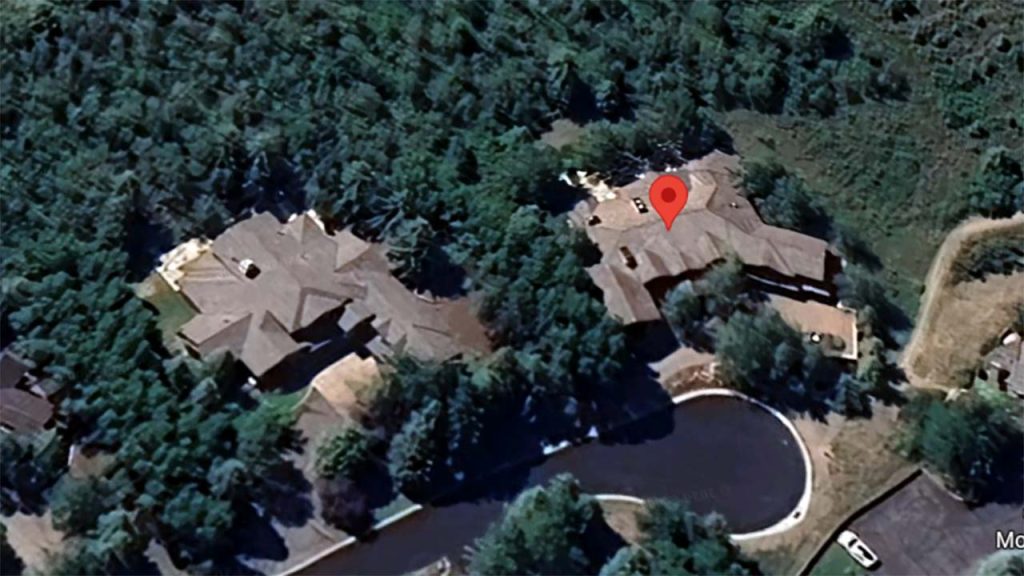 Aerial view of two large houses surrounded by dense green trees. A pin marks one house, suggesting a location point. The scene conveys a serene, secluded atmosphere.