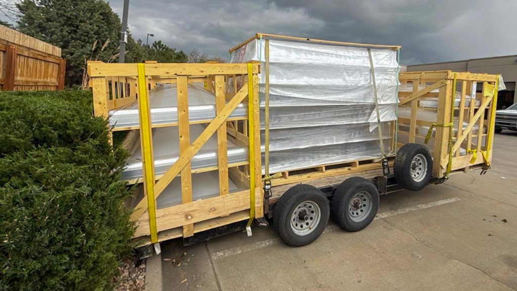 A large trailer with a wooden frame carrying long metal skylight curbs and smoke vent domes is parked beside bushes. The overcast sky sets a gloomy tone to the industrial scene.