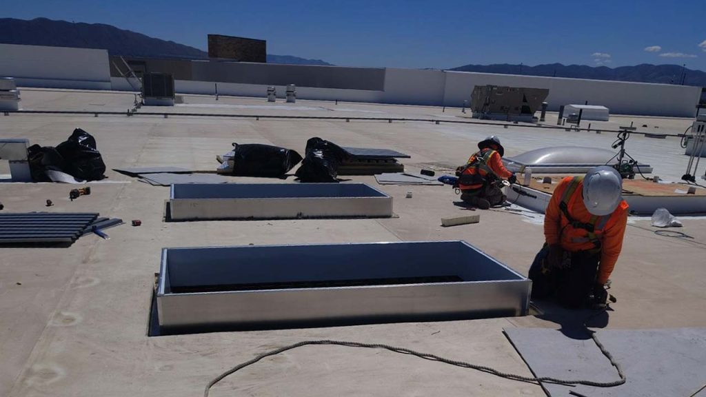 Two construction workers in orange safety vests and helmets work on installing smoke vent skylights on a large, flat rooftop under a clear blue sky.
