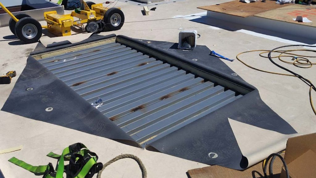 A rooftop construction scene shows a metal vent hole covered by black material, surrounded by tools, cables, and a yellow machine under bright sunlight.