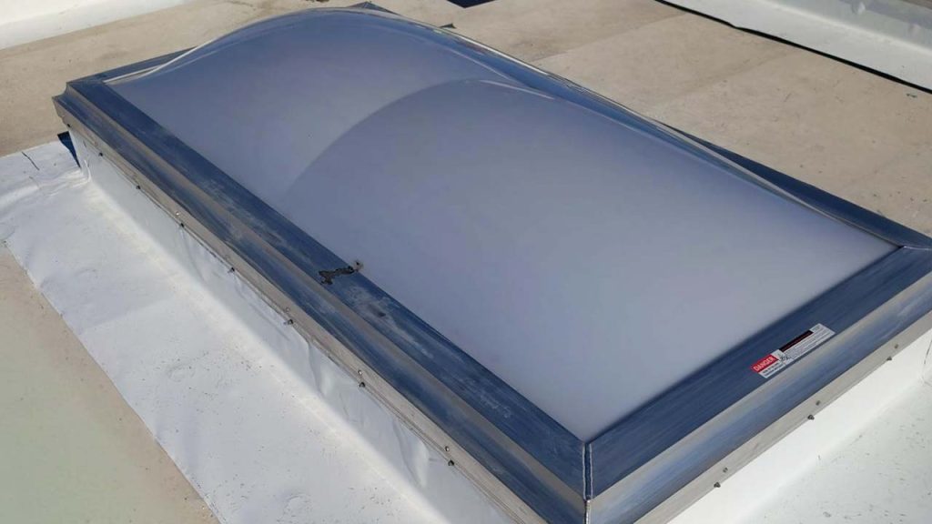 A domed smoke vent skylight with a white, translucent surface is set in a metallic frame on a flat rooftop. Sunlight casts slight shadows, creating a calm atmosphere.