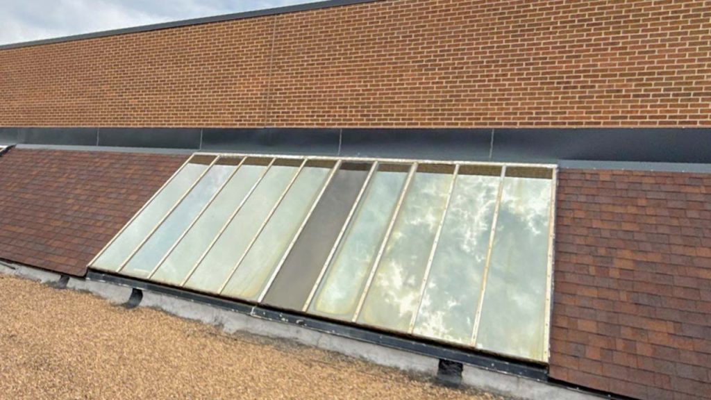 Single slope skylight, framed by a brown shingled roof, reflects a cloudy sky. Brick wall background adds contrast to the scene.