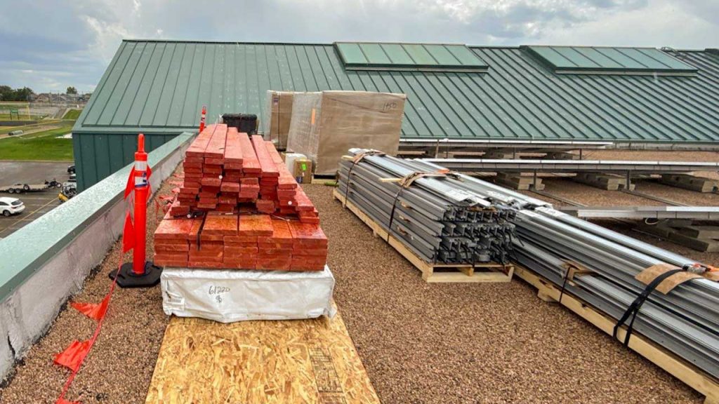 Stacks of red lumber and metal beams are neatly arranged on a rooftop under a cloudy sky. The scene conveys a sense of construction and organization.