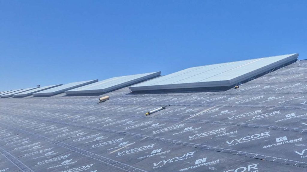 A sloped roof under bright blue sky, covered with black Vycor brand underlayment. Several skylights are visible, and roofing tools are scattered around.