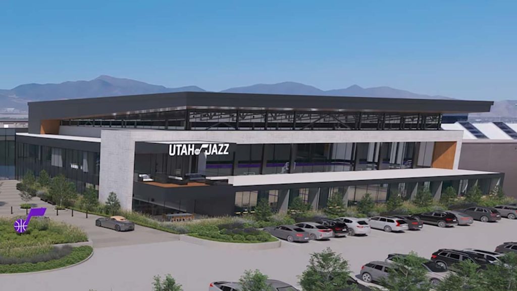 Modern building with "Utah Jazz" sign, large windows, surrounded by parked cars and greenery. Mountain range in the background under a clear sky.