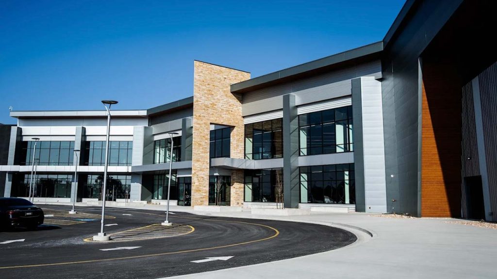 Utah Mammoth Training Facility building with large glass windows, stone accents, and sleek metal detailing. Clear blue sky above and an empty curved driveway in front.