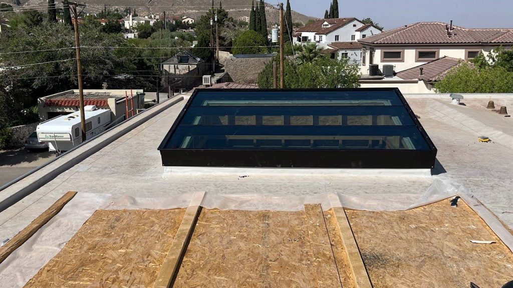 A rectangular glass skylight is installed on a flat rooftop, surrounded by residential buildings and trees under a clear blue sky. The scene feels calm and open.