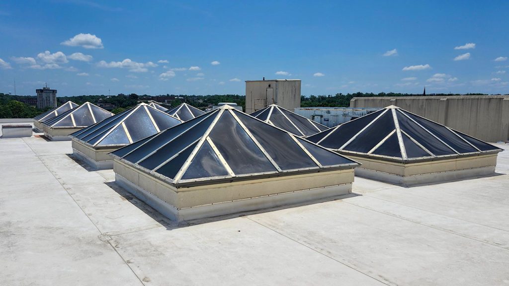 Pyramid-shaped skylights on a flat rooftop under a clear blue sky with scattered clouds. The scene conveys a sense of openness and modern architecture.