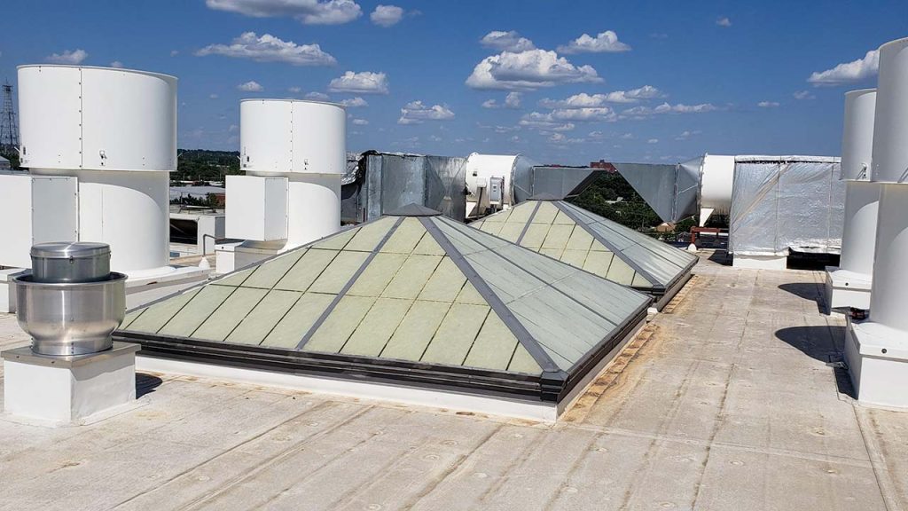 Rooftop with white HVAC units, large green-tinted pyramid skylights, and metallic ductwork under a bright blue sky with scattered fluffy clouds.