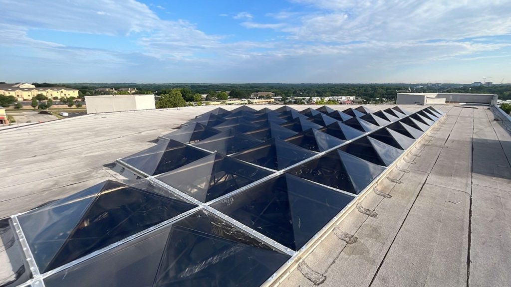 A rooftop with a large cluster of dark, polycarbonate, pyramid skylights under a partly cloudy blue sky. The urban landscape is visible in the distance.