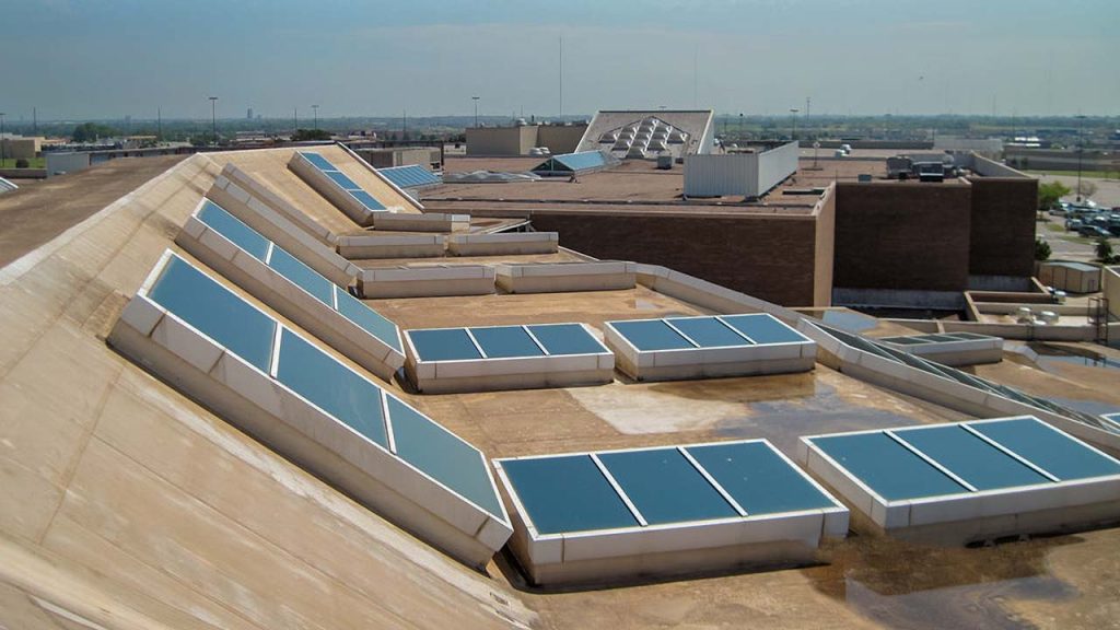 A flat rooftop with multiple rows of skylights angled towards the sky, set against an industrial background with clear skies, suggesting sustainability.