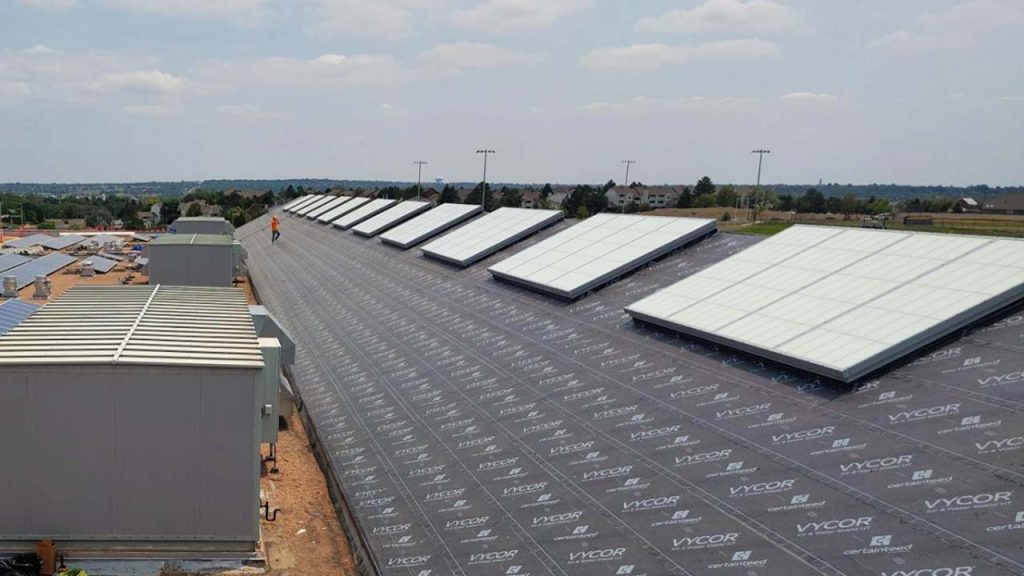 Wide aerial view of a school flat roof with a worker standing beside a row of large, rectangular translucent GridSpan skylight panels installed over underlayment.