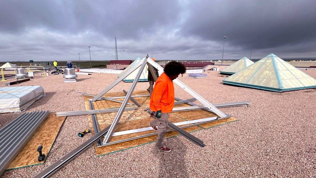 Commercial skylight installer assembling metal framing for a VELUX GridSpan pyramid skylight on a gravel rooftop at Limon Correctional Facility.