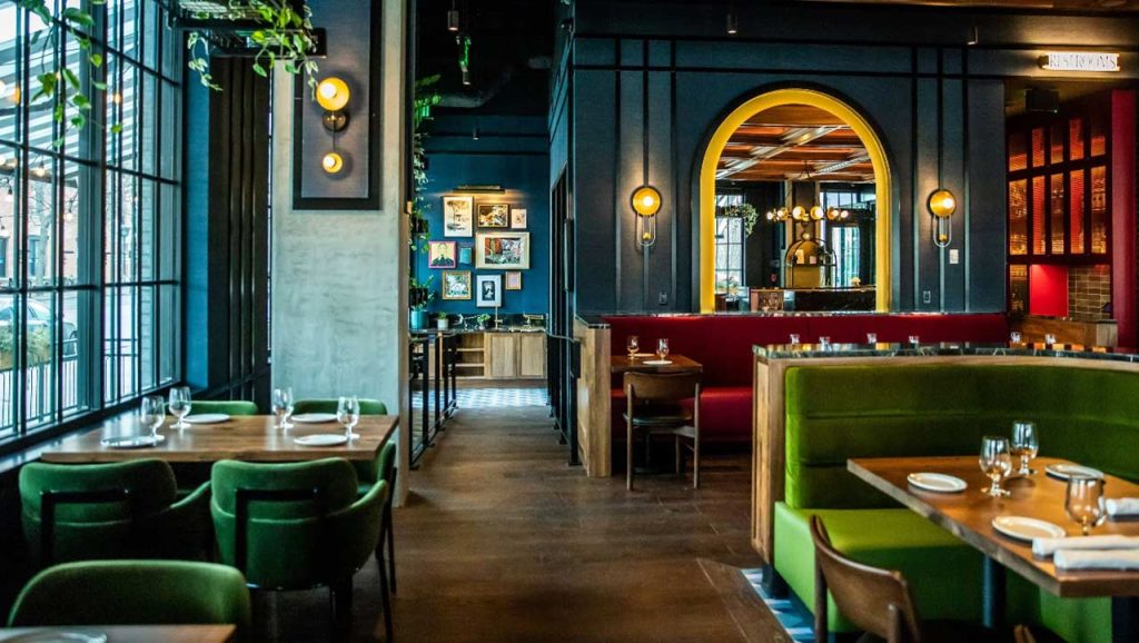 Interior view of a stylish restaurant dining room featuring dark blue walls, green upholstered booth seating and dining chairs, warm wood tables, set places, and decorative archways with warm lighting.